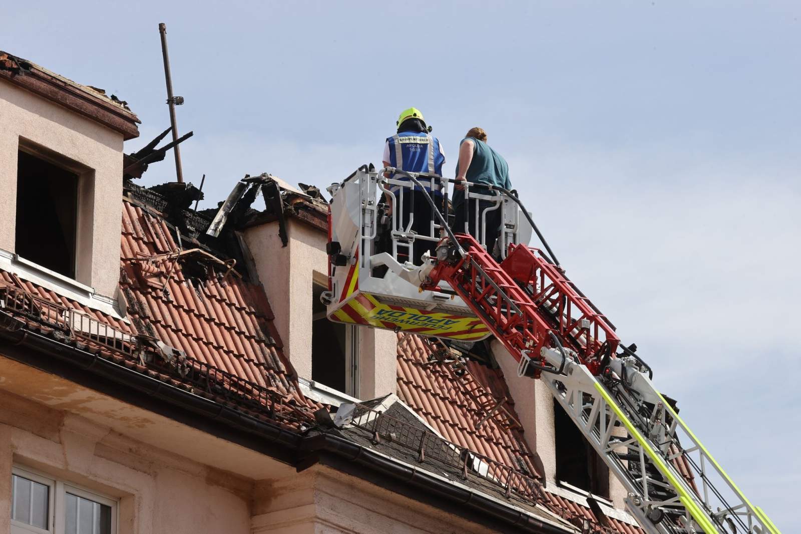 Feuerwehrleute und Statiker untersuchen das Dach eines Mehrfamilienhauses von einer Drehleiter aus.