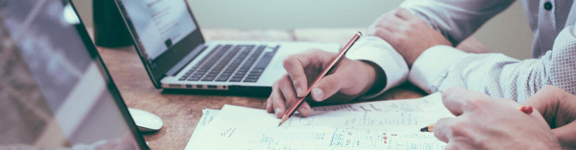 Zwei Personen sitzen am Tisch mit zwei Laptops vor ihnen. Vor ihnen liegt noch Papier, © Shutterstock