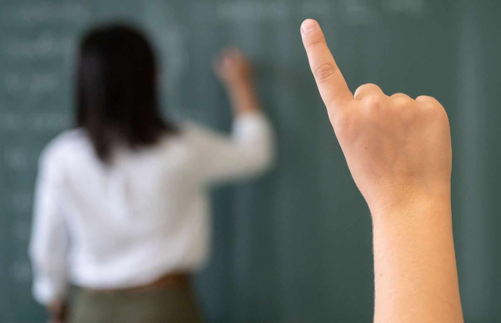 Ein Schüler meldet sich, während die Lehrerin an die Tafel schreibt., © Marijan Murat/dpa/Symbolbild