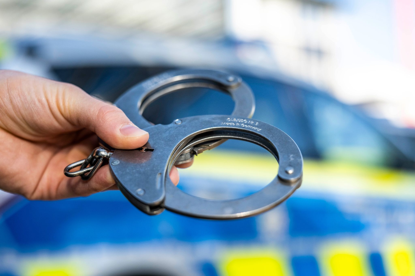 Eine Hand hält Handschellen vor einen Streifenwagen der Polizei., © David Inderlied/dpa/Illustration