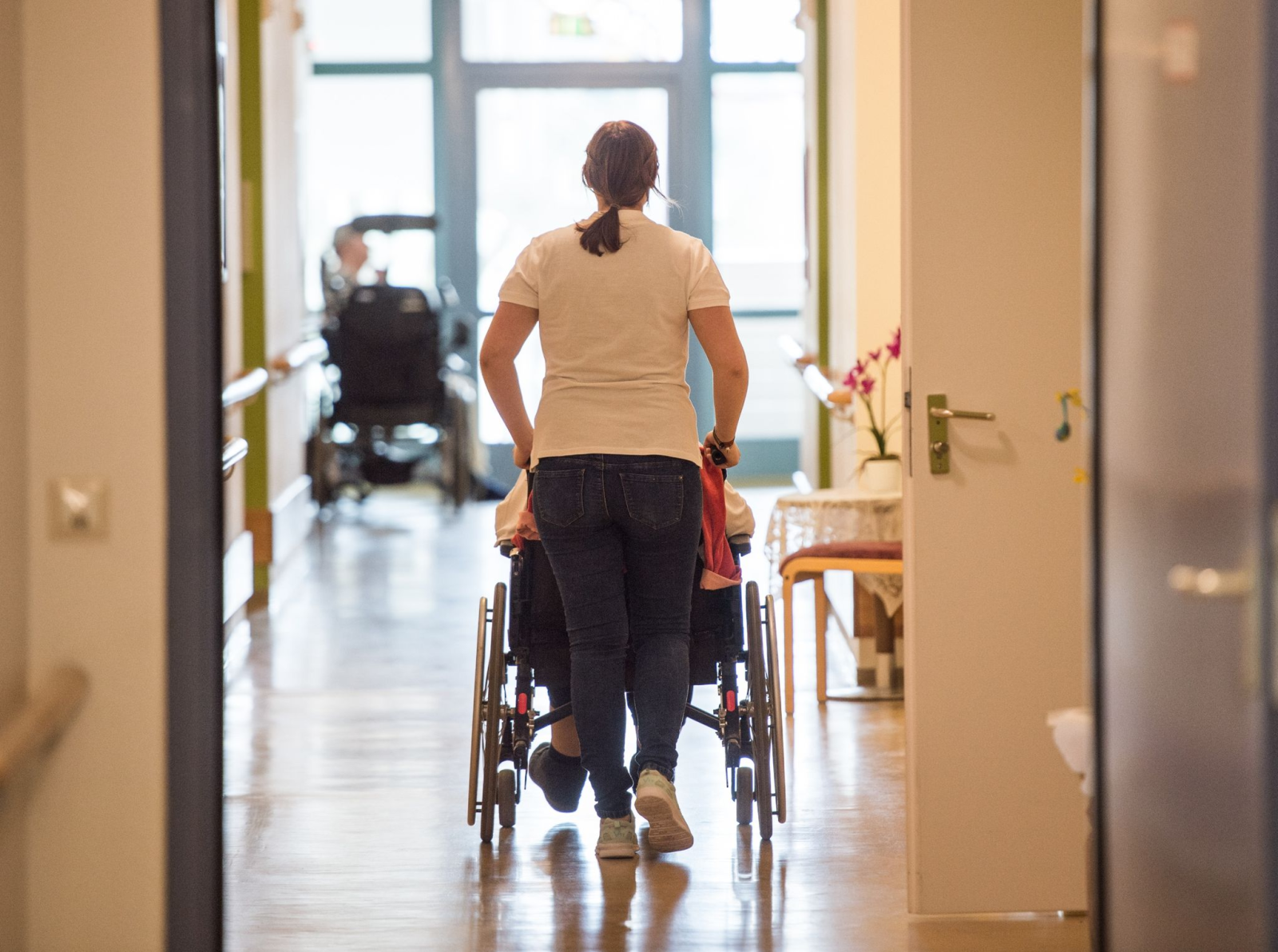 Eine Pflegerin schiebt eine ältere Frau im Rollstuhl durch einen Flur im Seniorenzentrum.