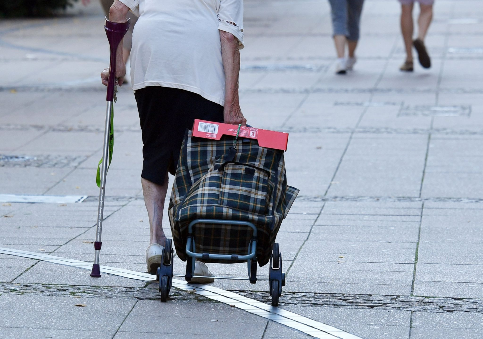 Eine alte Frau mit einer Krücke und Einkaufstrolley geht durch eine Fußgängerstraße.
