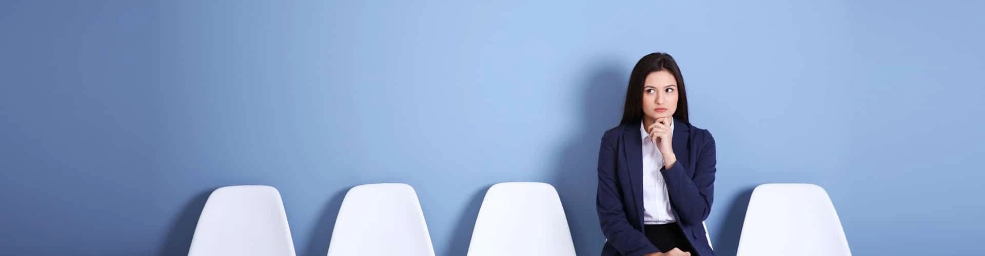 Frau sitzt auf einem Stuhl, neben ihr vier weiße leere Stühle. Sie hat ihre Hand am Kinn, © Shutterstock Frau sitzt auf einem Stuhl, neben ihr vier weiße leere Stühle. Sie hat ihre Hand am Kinn, © Shutterstock