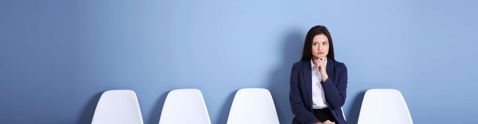 Frau sitzt auf einem Stuhl, neben ihr vier weiße leere Stühle. Sie hat ihre Hand am Kinn, © Shutterstock