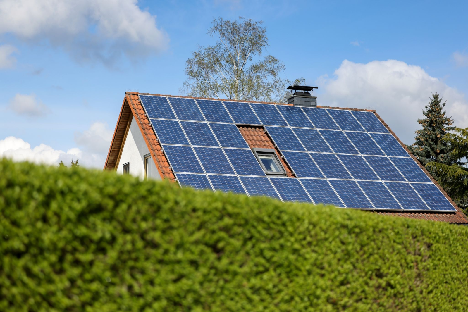 Ein Einfamilienhaus mit Solarpaneelen auf dem Dach steht hinter einer grünen Hecke.